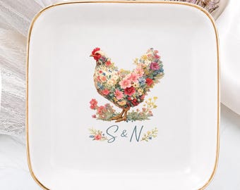 Puede incluir: Un plato cuadrado de cerámica blanca con un borde dorado. El centro presenta una ilustración colorida de una gallina compuesta por varias flores, situada sobre el texto azul S & N, flanqueado por pequeñas ramas de flores.