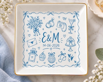 Può includere: Un piattino quadrato in ceramica bianca con bordo dorato, decorato con illustrazioni a linee blu di icone a tema matrimonio, tra cui un bouquet, anelli, fotocamera, torta e palla da discoteca, con il testo centrale E & M e 14-06-2026.