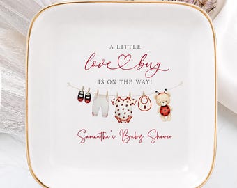Può includere: Un piattino quadrato in ceramica bianca con bordo dorato. Al centro è presente il testo A LITTLE love bug IS ON THE WAY! sopra un'illustrazione di vestiti per neonati su un filo da bucato e il testo Samantha's Baby Shower in rosso.