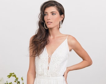May include: A woman wearing a white lace dress with a deep V-neckline. She has long brown hair and is wearing large pearl hoop earrings.