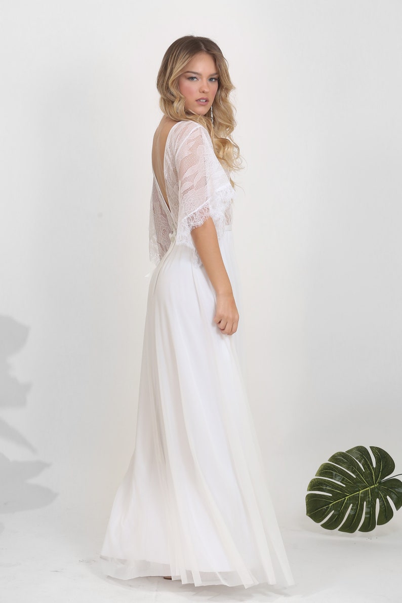 May include: A white lace wedding dress with a flowing skirt and a low back. The dress has a delicate lace overlay and a simple, elegant design.