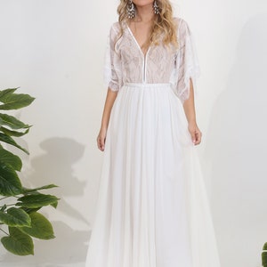 May include: A white lace wedding dress with a V-neckline and a flowing skirt. The dress has a sheer overlay and a delicate lace pattern.