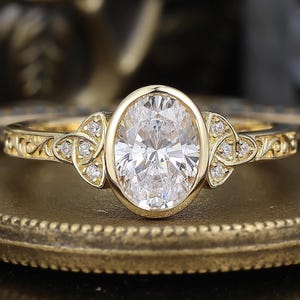 Op de afbeelding: Een gouden verlovingsring met een ovaal geslepen diamant als middelpunt. De ring heeft kleinere diamanten aan weerszijden van de ovaal en ingewikkelde filigraan details langs de band. De ring ligt op een goudkleurig oppervlak.