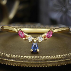 May include: A gold-colored ring with a curved band. The ring features pink and white gemstones, and a teardrop-shaped blue gemstone. The ring is set on a gold-colored surface.