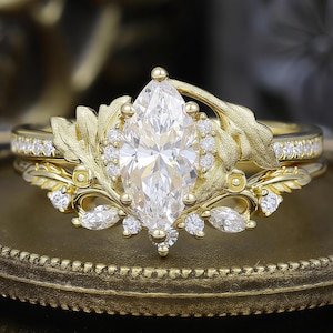 May include: A gold engagement ring set with a marquise-cut diamond center stone. The ring features leaf and vine detailing with smaller accent diamonds. The band is also adorned with small diamonds, creating a sparkling effect.