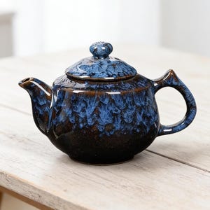 Può includere: Una teiera in ceramica con smalto blu e marrone. La teiera ha un corpo arrotondato, un beccuccio curvo e un manico ad anello. Il coperchio è sormontato da un piccolo pomello. La teiera è appoggiata su una superficie di legno chiaro.