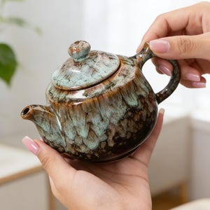 Puede incluir: Una pequeña tetera de cerámica con tapa, asa y boquilla. La tetera tiene un esmalte moteado en tonos marrones, verdes y azules. La tetera se sostiene con dos manos.