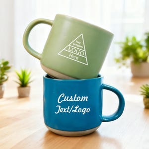 Puede incluir: Dos tazas de cerámica apiladas. La taza superior es verde con un logotipo blanco y el texto "Your Company LOGO Here". La taza inferior es azul con el texto "Custom Text/Logo". Ambas tazas tienen una base clara y un asa curva.