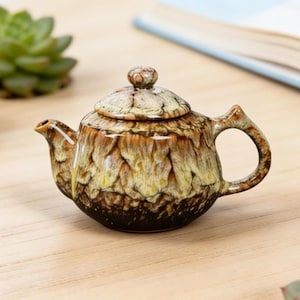 May include: A small ceramic teapot with a lid. The teapot features a textured, layered design in shades of brown, tan, and yellow. The handle and spout are curved. The teapot is on a wooden surface.