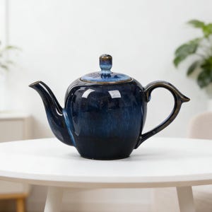 May include: A dark blue glazed ceramic teapot with a lid. The teapot has a curved spout and handle, and is placed on a white table. This teapot is a stylish piece of kitchenware.