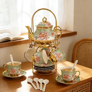 Może przedstawiać: Zestaw do herbaty w stylu vintage ze złotym metalowym stojakiem. Zestaw zawiera czajniczek, filiżanki, spodki i łyżeczki. Zestaw do herbaty ma jasnozielone tło z różowymi kwiatowymi wzorami i złotymi obwódkami. Zestaw do herbaty jest prezentowany na drewnianym stole.