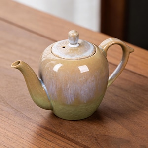 Könnte beinhalten: Eine Keramik-Teekanne mit rundem Körper, Ausguss und Henkel. Die Teekanne hat einen Deckel mit einem kleinen Knopf. Der Farbverlauf geht von einem hellen Grün am Boden zu einem Goldbraun mit weißen und blauen Akzenten über.