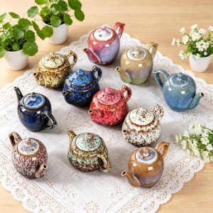 May include: A collection of small, ceramic teapots in various colors, including blue, red, brown, and green. Each teapot has a unique glaze and design, with a spout, handle, and lid. The teapots are arranged on a white lace tablecloth.