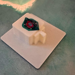 Op de afbeelding: Een wit, vierkant object met een kleiner, rechthoekig blok erbovenop. Op het bovenste blok is een groene en rode sticker aangebracht. Het object staat op een groter, wit vierkant, tegen een turquoise achtergrond.