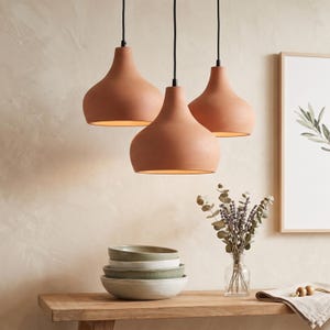 Puede incluir: Tres lámparas colgantes de color terracota con pantallas lisas en forma de campana cuelgan de cables negros. Debajo, una pila de cuatro cuencos de cerámica en tonos verde apagado y blanco roto sobre una superficie de madera junto a un florero.