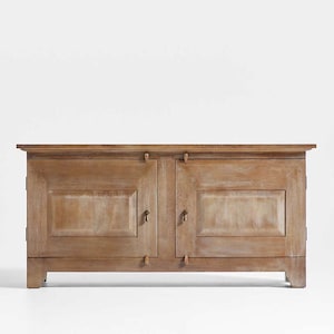 May include: A light brown wooden cabinet with two doors and small wooden handles. The cabinet has a rectangular shape and a slightly distressed finish, with visible wood grain. The doors have recessed panels, and the cabinet sits on short, square legs.