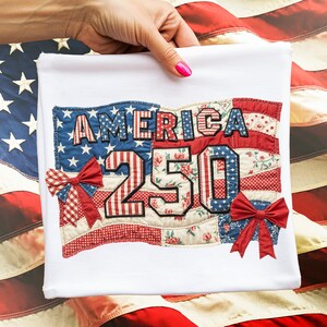 Op de afbeelding: Wit T-shirt met een patriottisch ontwerp. Het ontwerp toont het woord "AMERICA" en het nummer "250" in patchwork-stijl. Het ontwerp bevat rode, witte en blauwe stoffen, waaronder bloemen- en sterrenpatronen, en is geaccentueerd met rode strikken.
