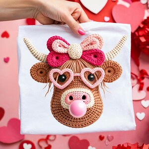 Op de afbeelding: Wit T-shirt met een gehaakt Highland koeienontwerp. De koe heeft hartvormige brillen, een roze neus en een strik. De hoorns zijn crèmekleurig en het gezicht van de koe is bruin. Het ontwerp is gecentreerd op het shirt.