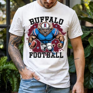 May include: White t-shirt featuring a muscular buffalo-themed football player graphic. The design includes the text "BUFFALO FOOTBALL" in bold, dark lettering. The player is depicted in a blue and white uniform, holding a football.