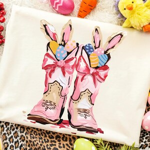 May include: A cream-colored Easter-themed design featuring pink cowboy boots with bunny ears and colorful Easter eggs. The boots have pink bows and are decorated with a floral design. The image is surrounded by Easter decorations.