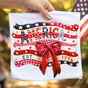 Może przedstawiać: Biały ręcznik dekoracyjny z patriotycznym wzorem. Ręcznik zawiera napis "AMERICA" w kolorowej czcionce, czerwoną kokardę i rok "1776". Projekt zawiera elementy czerwone, białe i niebieskie, gwiazdy i paski.