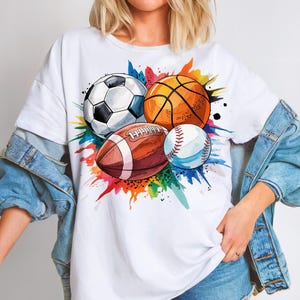 Puede incluir: Camiseta blanca con un gráfico colorido de un balón de fútbol, baloncesto, fútbol americano y béisbol. Las pelotas deportivas están rodeadas de vibrantes salpicaduras de pintura en tonos azules, naranjas, verdes y rojos. La camiseta es holgada.