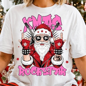 Puede incluir: Camiseta blanca con un gráfico de Papá Noel con una chaqueta de cuero roja, gafas de sol y un gorro de Papá Noel. El diseño incluye el texto "XMAS ROCKSTAR" en rosa, con guitarras y estrellas.