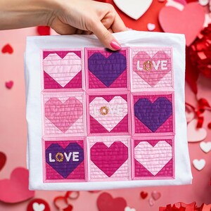 May include: White t-shirt featuring a patchwork design of nine hearts in shades of pink and purple. The word "LOVE" is embroidered on three of the hearts. The design is square and centered on the shirt, perfect for Valentine's Day.