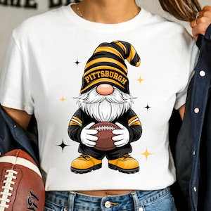 May include: White t-shirt featuring a cartoon gnome wearing a black and gold football uniform and a hat that says "Pittsburgh". The gnome is holding a brown football. The design includes star accents.