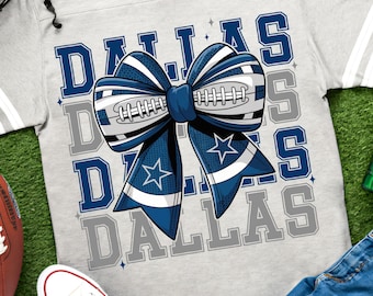 May include: A light gray V-neck t-shirt featuring a large graphic of a blue and white bow designed with football laces and star patterns. The word DALLAS is printed repeatedly in the background in blue and gray block lettering on the front.