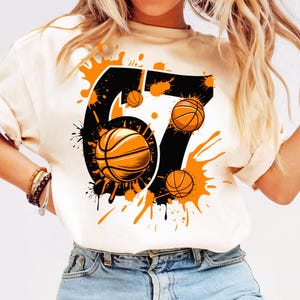May include: Cream-colored t-shirt featuring a black number 67 with orange basketballs and paint splatter design. The basketballs are orange with black lines. The shirt is a casual style.