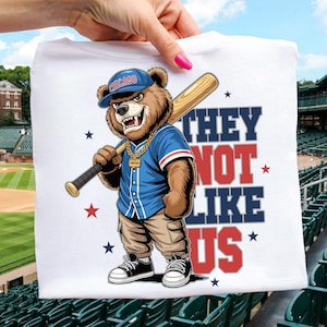 May include: White t-shirt with a cartoon bear wearing a baseball cap with "Chicago" on it, holding a baseball bat. The bear is dressed in a blue jersey and khaki trousers. The text "THEY NOT LIKE US" is printed on the shirt.