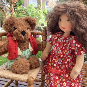 May include: A brown teddy bear wearing a red knitted scarf and a green knitted waistcoat sits on a wicker chair. A doll with long brown hair and a red floral dress stands next to the chair.