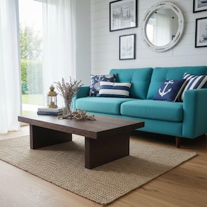 Peut inclure: Un canapé bleu sarcelle avec trois coussins décoratifs à motifs d'ancres et de rayures, placé derrière une table basse en bois brun foncé. La table supporte une petite lanterne, un vase en verre avec des fleurs séchées et des livres.