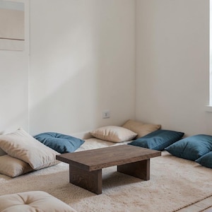 Könnte beinhalten: Ein rechteckiger Couchtisch aus dunkelbraunem Holz steht auf einem cremefarbenen Teppich. Mehrere quadratische Kissen in Blau- und Beigetönen umgeben den Tisch und schaffen eine gemütliche Sitzecke.