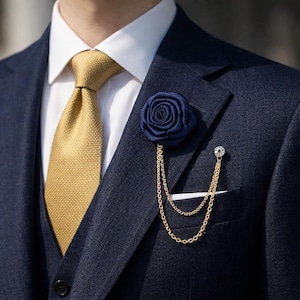 May include: A navy blue rose lapel pin with a gold chain, worn on a navy suit. The suit features a gold tie and a white shirt. A small, round, jeweled pin secures the chain, adding a touch of elegance.