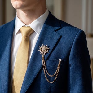 May include: A gold-colored brooch in the shape of a starburst with a central clear gem, attached to a double chain. The brooch is pinned to a blue blazer, paired with a gold tie and white shirt. A small, clear gem adorns the blazer pocket.