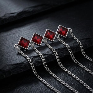 May include: A silver-toned chain bracelet featuring four square, ruby-red gemstones. Each gem is set in a silver frame and connected to a delicate chain. The bracelet is displayed on a dark, textured surface, highlighting the contrast between the bright gems and the background.