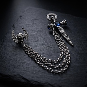 May include: Silver-toned brooch featuring a skull with wings, a dagger with blue gemstones, and multiple chain strands. The brooch has a gothic or punk aesthetic, with intricate detailing and a dark, edgy style.