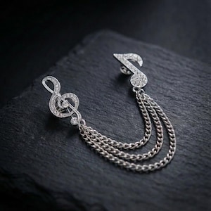 Puede incluir: Broche plateado con forma de clave de sol y una nota musical, ambos adornados con cristales transparentes. El broche tiene tres cadenas colgantes. La joya está sobre un fondo oscuro y texturizado.