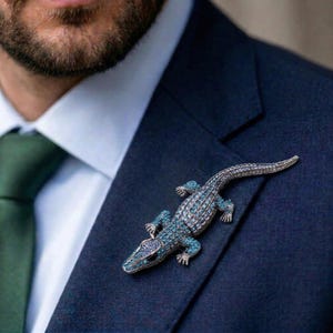Puede incluir: Broche de lagarto enjoyado en tonos azules y plateados, fijado a una chaqueta de traje azul marino. La broche presenta detalles intrincados y es un accesorio llamativo. El cuerpo del lagarto está cubierto de pequeñas piedras brillantes.