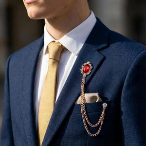 May include: A gold-toned brooch with a large red gemstone and a chain, attached to a navy blue suit. The brooch is worn on the lapel, with a gold tie and pocket square visible.