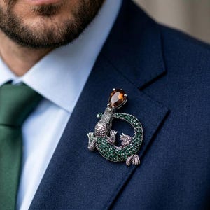 May include: A silver alligator brooch with green and clear crystal accents and a teardrop-shaped amber stone. The brooch is pinned to a navy blue suit jacket, with a green tie visible.