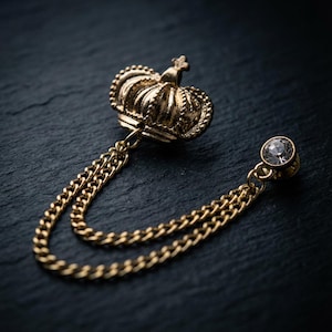 May include: A gold-colored crown lapel pin with a chain and a crystal accent. The crown is detailed with intricate designs and a small cross on top. The chain connects the crown to a crystal stud. The pin is set against a dark background.
