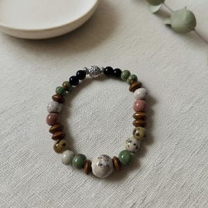 May include: A beaded bracelet featuring a variety of earth-toned beads. The bracelet includes round beads in shades of brown, green, white, and pink, interspersed with smaller wooden and black beads. A silver-colored clasp secures the bracelet.