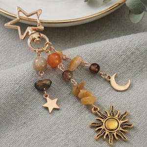 May include: Gold-toned keychain featuring a star-shaped clasp and dangling charms. The charms include a star, a crescent moon, and a sunburst, along with various beads in shades of brown, orange, and yellow. The keychain is designed with a celestial theme.