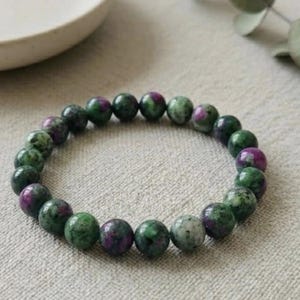 May include: A beaded bracelet featuring round, polished gemstones. The beads are a mix of green and deep pink, strung together to form a circular shape. The bracelet is displayed on a neutral-toned surface.