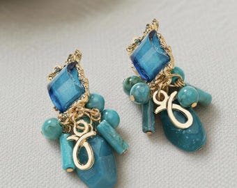 May include: A pair of turquoise and gold earrings. Each earring features a large, square, blue gemstone set in a gold filigree frame. Below the gemstone are turquoise beads and a gold charm.