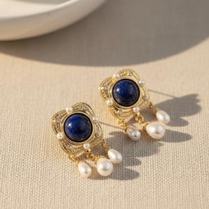 以下が含まれることがあります： 正方形のデザインのゴールドトーンのイヤリングのペア。各イヤリングは、中央に大きな丸い濃い青色の石があり、小さな白い真珠と透明なクリスタルに囲まれています。3つのぶら下がっている白い涙型の真珠が下からぶら下がっています。
