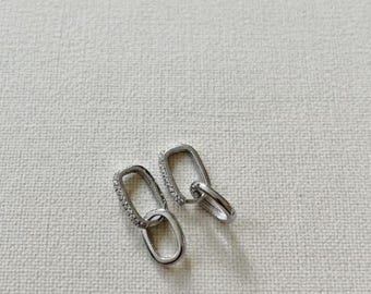 May include: A pair of silver-toned earrings. One earring features a rectangular loop with a row of small, clear stones. The other earring has a similar rectangular loop connected to a smaller, plain loop. The earrings are displayed on a textured, off-white surface.
